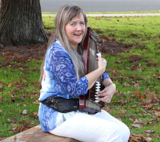 Jacqui sitting on a bench playing her autoharp